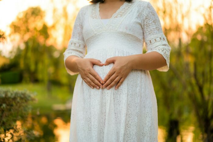Pregnant woman in white dress forming heart shape on belly in outdoor wedding setting