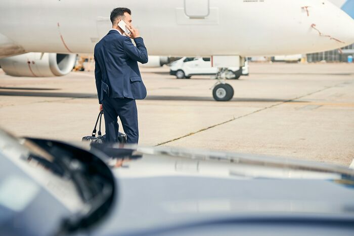 A man in a suit talking on the phone, holding a bag, standing on an airport tarmac near a private jet, representing rich people's silent luxuries.