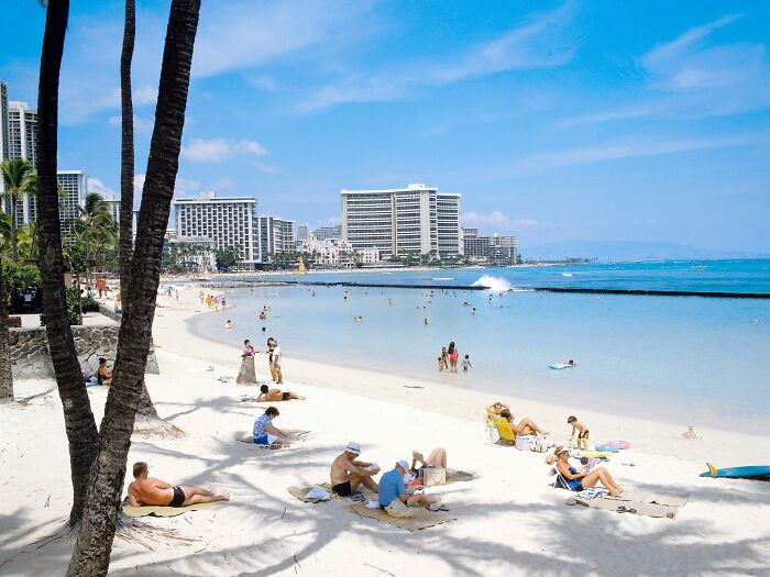 Tourists relaxing on a sunny beach in front of hotels and blue ocean, palm trees in the foreground.