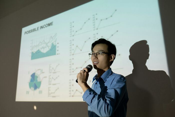 A man in glasses speaks into a microphone in front of a projected graph display, a cringe moment for second-hand embarrassment.
