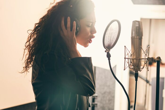 A woman with long curly hair and headphones sings into a studio microphone, representing silent luxuries.