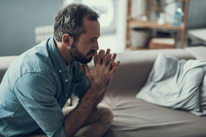 A bearded man in a denim shirt sits on a sofa, looking pensive with clasped hands, reflecting on why men are suffering.