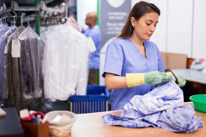 A woman in scrubs and yellow-green gloves brushes a plaid shirt at a dry cleaner. Rich people's silent luxuries.