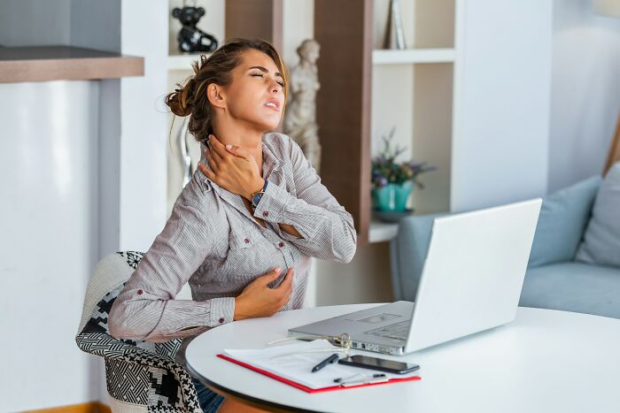 A woman holds her throat and chest in discomfort at a table with a laptop, reflecting concerning facts.