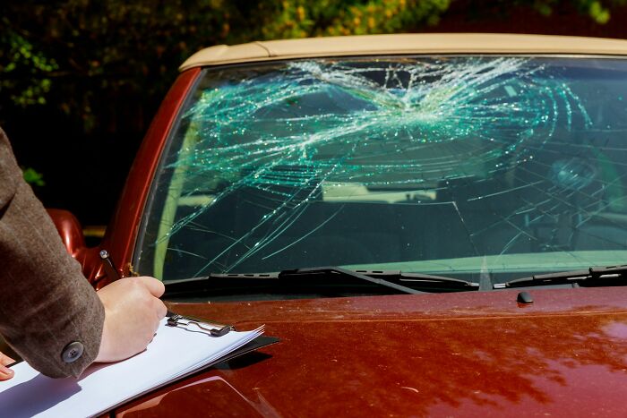 Person writing on a clipboard in front of a car with a shattered windshield, reflecting disturbing d***h stories.