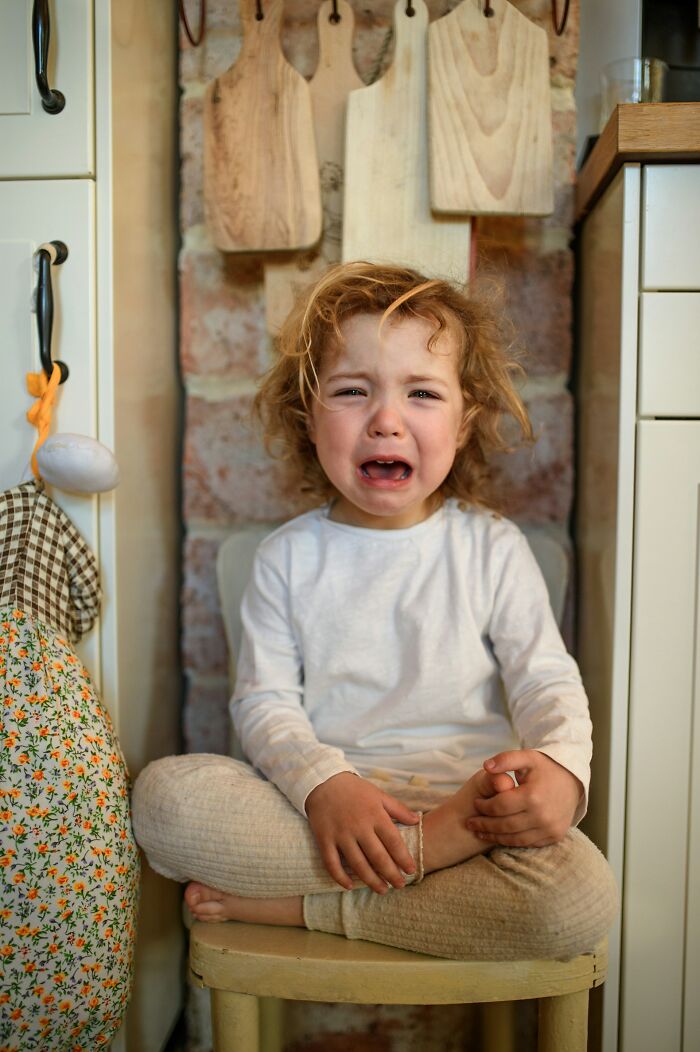 A young, curly-haired child in a white shirt and beige pants sits crying on a stool, expressing deeply immoral emotions.