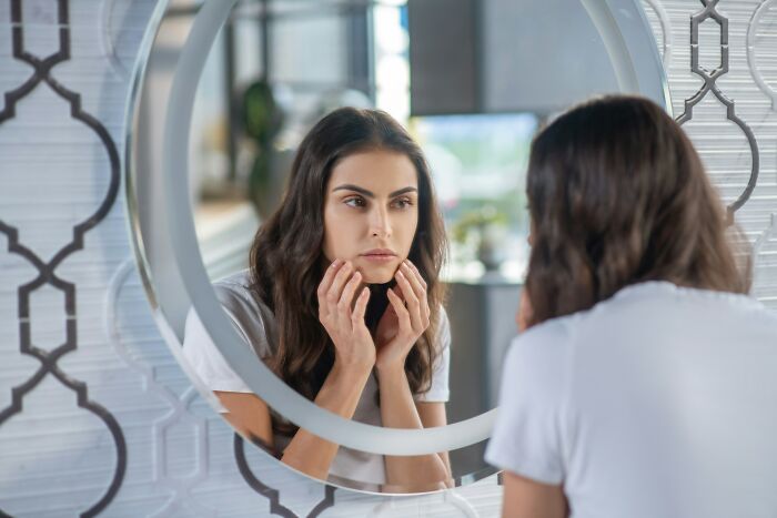 Woman looking closely at her skin in a round mirror, concerned. Focus on routine cancer screening and health warning signs.