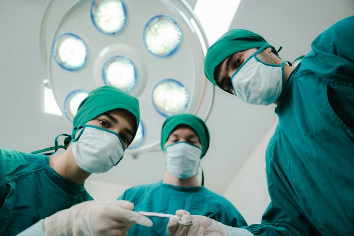 Doctors in surgical scrubs and masks look down, an operating room light above. Patients, how are you alive?