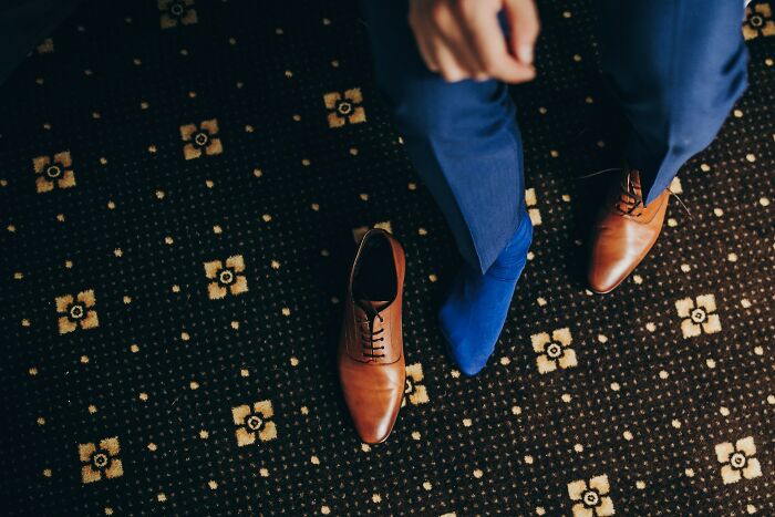 Overhead shot of a person in blue pants and socks, with a brown oxford shoe next to their foot. Rich people luxuries.