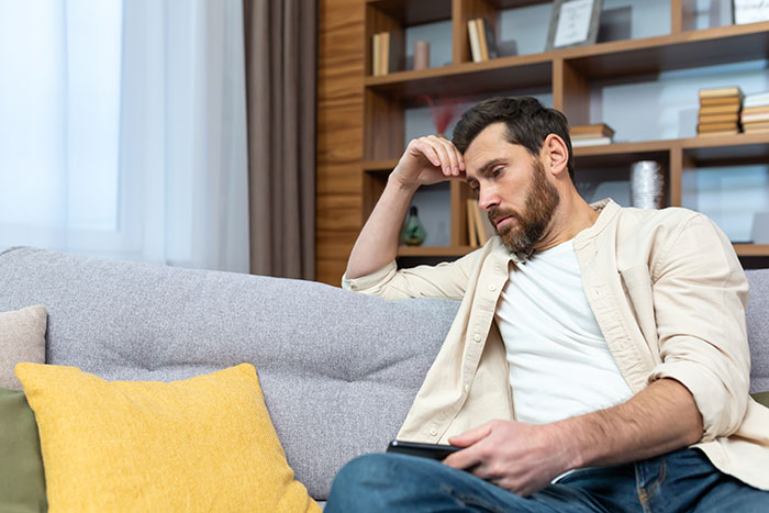 Man in beige shirt sitting on sofa looking worried holding phone Man in beige shirt sitting on sofa looking worried holding phone