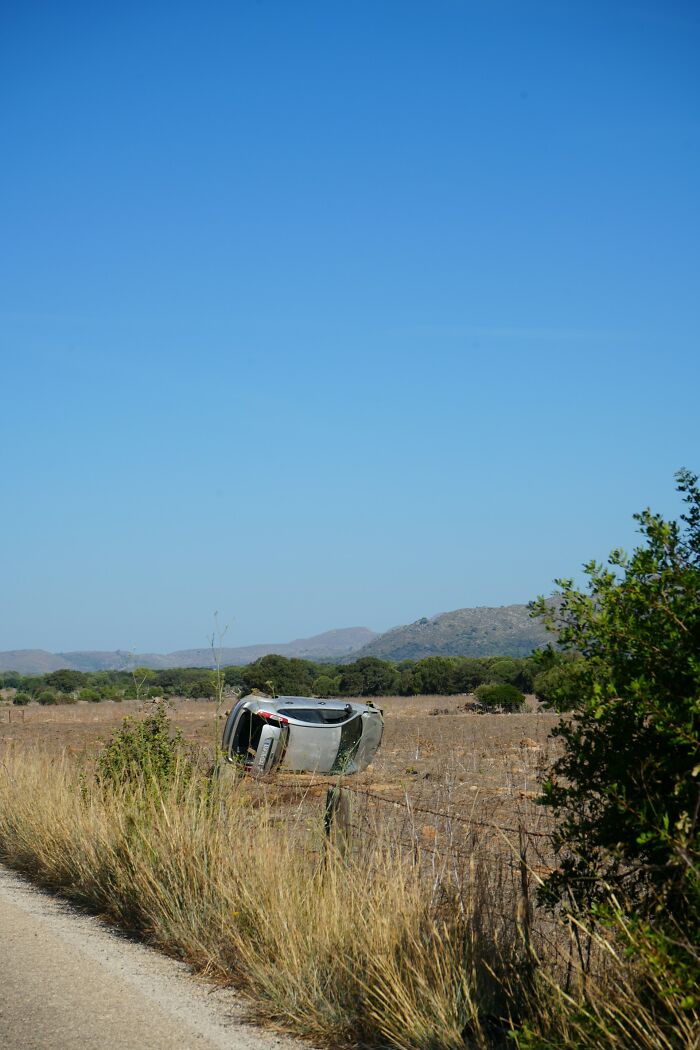 An overturned car, silver, rests on a fence in a dry, grassy field under a clear blue sky, hinting at disturbing d***h stories.
