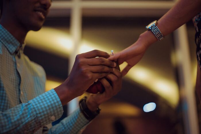 A man, wearing a blue checkered shirt, kneels to propose, placing an engagement ring on a woman's finger. Capturing a romantic story.