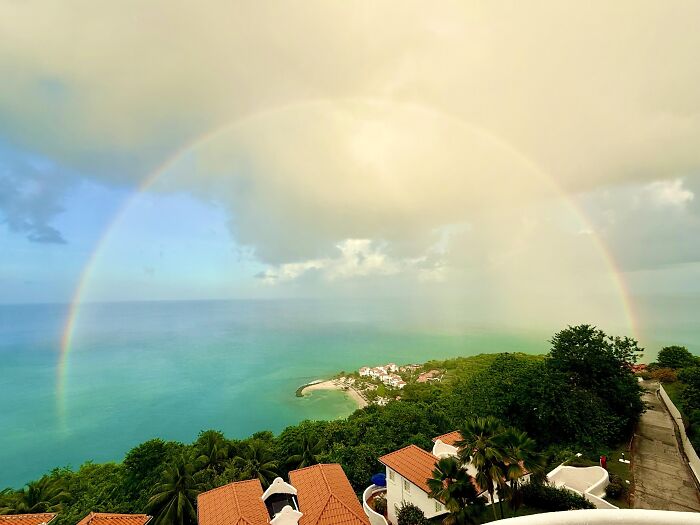 Full rainbow arching over coastal houses and ocean during partly cloudy day