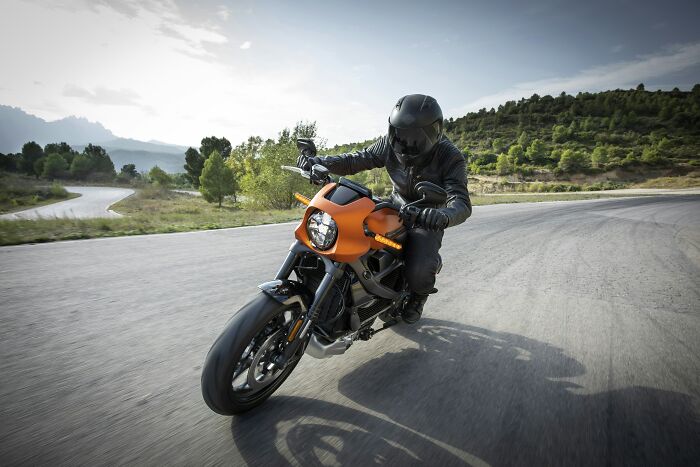 A motorcyclist in a black helmet and gear rides an orange bike on a winding road. Doctors may wonder, How are you alive?