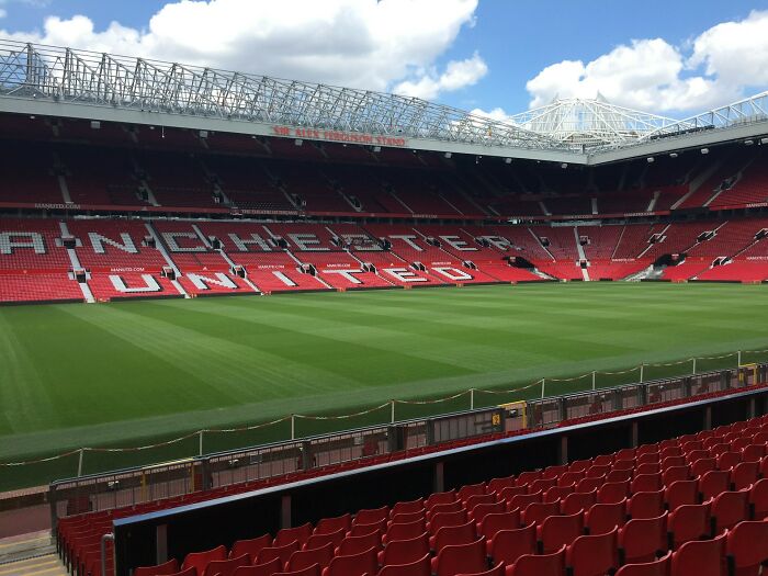 A vast football stadium, empty red seats surrounding a pristine green field, under a partly cloudy sky. Celebrity relatives.