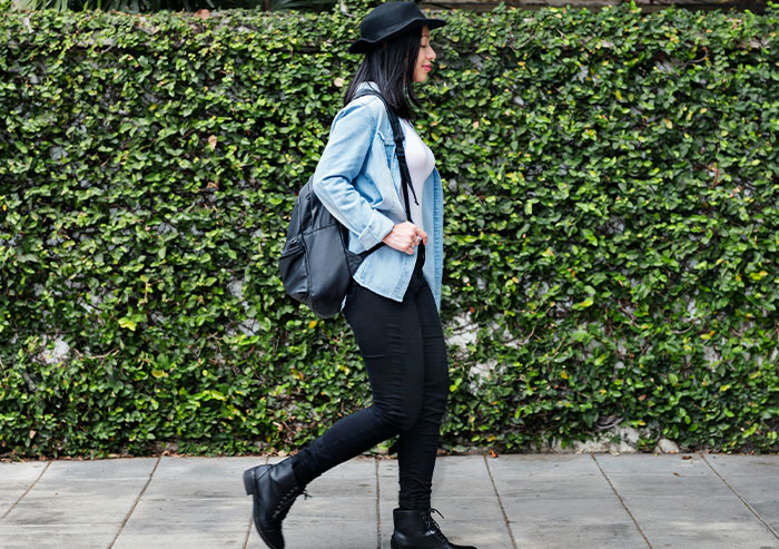 A young woman with a fedora, denim jacket, and backpack walks past a green ivy wall, reflecting high school crushes.