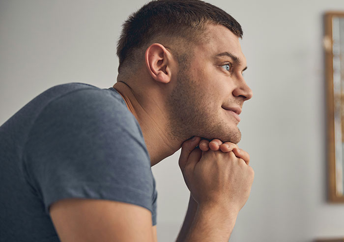 A man with a buzz cut and blue eyes, chin resting on clasped hands, smiling slightly. Reminiscing about high school crushes.