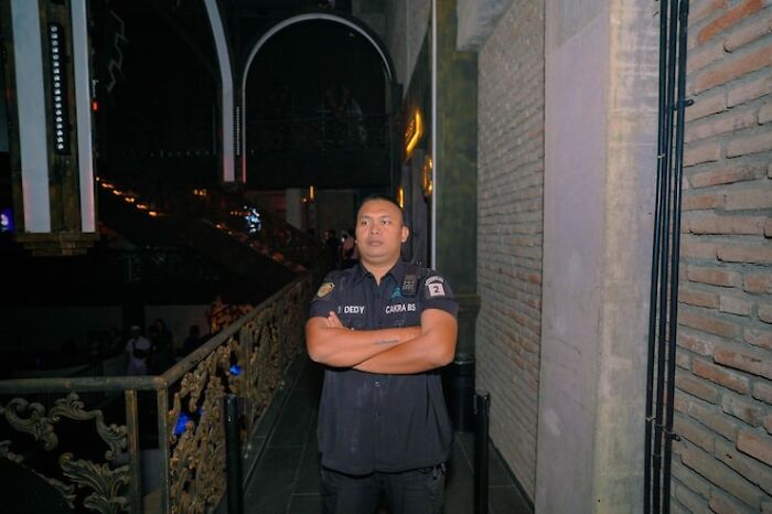 A security guard in a dark uniform stands with crossed arms in a dimly lit venue. He represents epic comebacks.