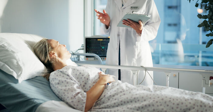 A doctor talks to a patient in a hospital bed. This image captures moments when doctors wonder "How the hell are you even alive right now?"