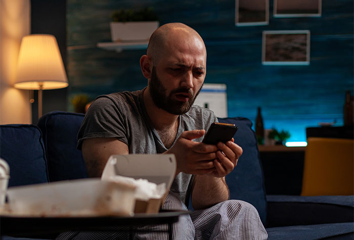 A man looks shocked, holding a phone, sitting on a couch. He finds his wife's photo on a friend's phone.