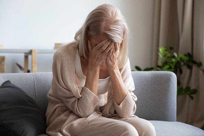 Older woman sitting on couch covering her face, showing sadness and heartbreak Older woman sitting on couch covering her face, showing sadness and heartbreak