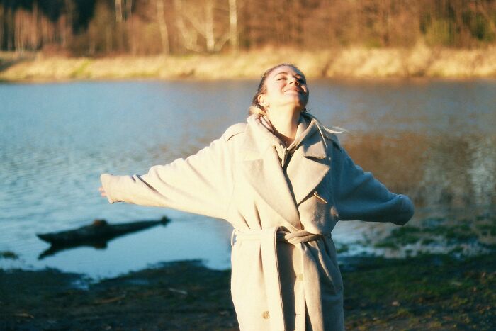 A woman in a beige coat with arms outstretched, eyes closed and head tilted back, enjoying nature near a lake. Cool facts on science.