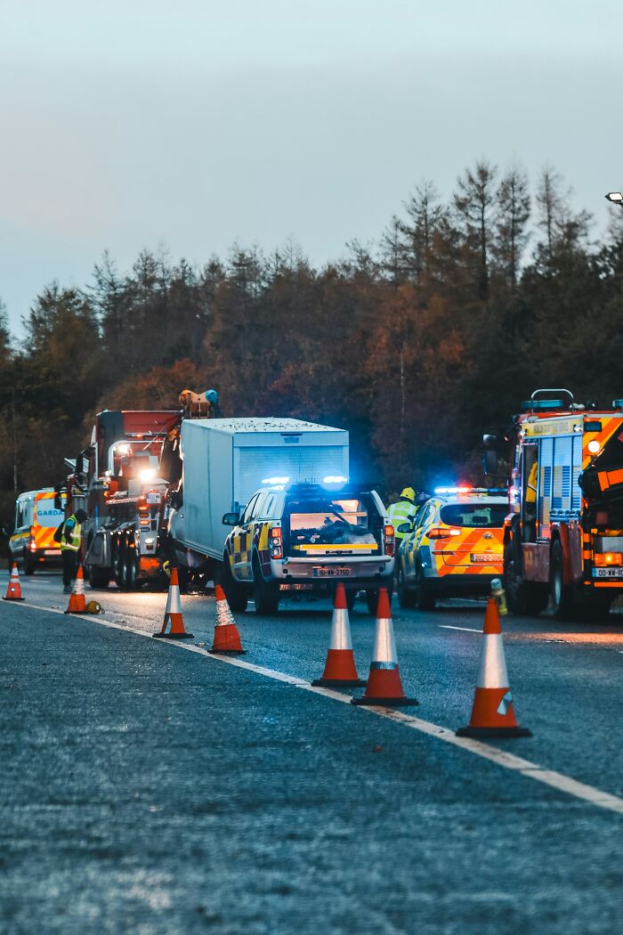 Emergency vehicles on a road at dusk, lights flashing, with traffic cones in the foreground. A creepy scene unfolding.