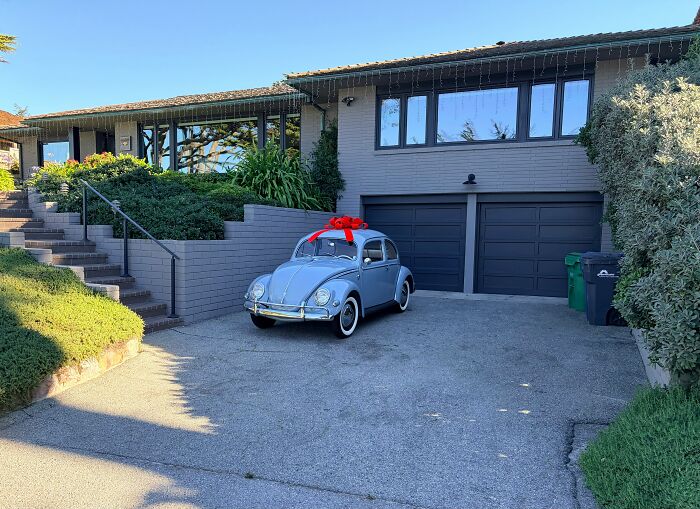 A classic light blue VW Beetle with a red bow sits in a driveway in front of a modern gray house, exposing privilege.
