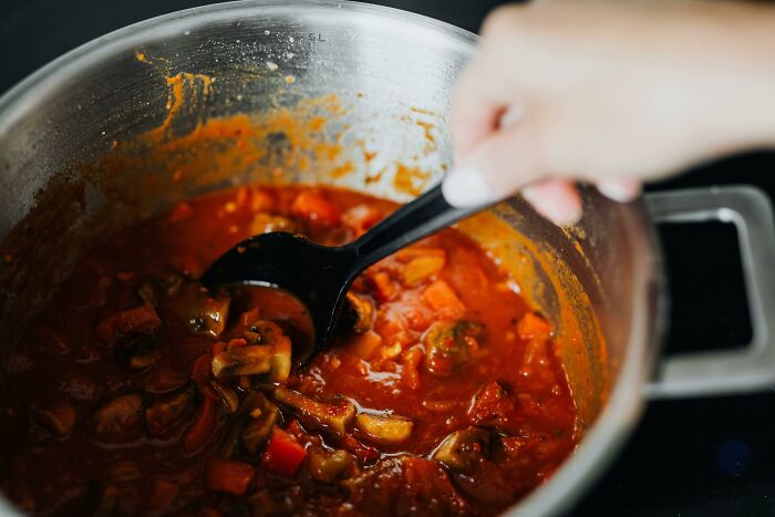Hand stirring a pot of simmering tomato sauce with mushrooms and vegetables, showing middle-class cooking habits.