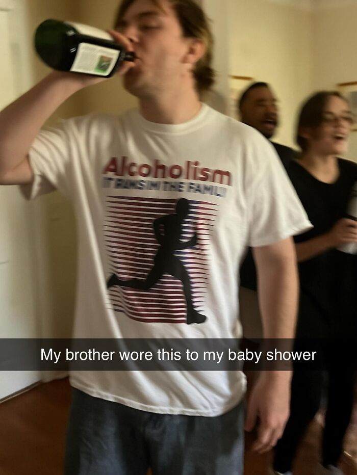 A man in a white T-shirt that says Alcoholism, It Runs In The Family, drinking from a bottle, at a baby shower.