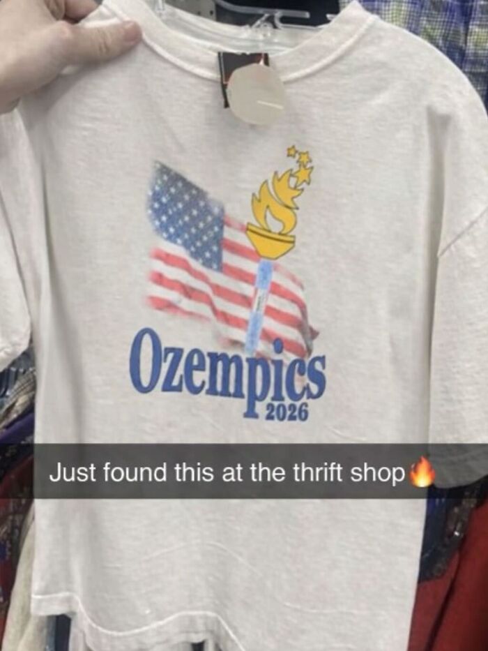 A hand holding up a white t-shirt with Ozempics 2026 printed below an American flag and a golden torch, an unhinged t-shirt find.