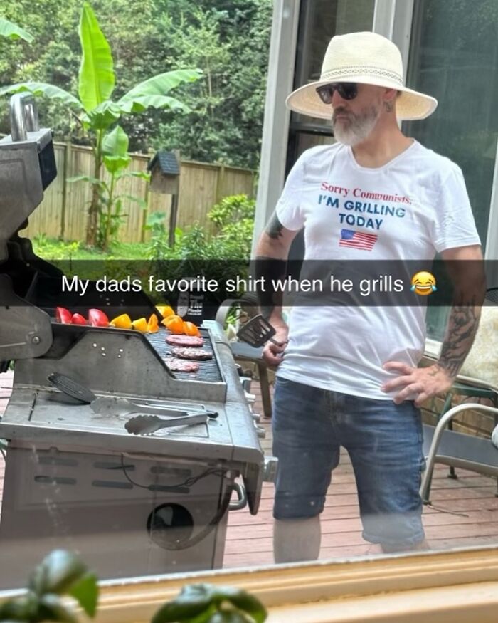 A man grilling outdoors, wearing a white t-shirt that says Sorry Communists, I'm grilling today with an American flag, showcasing bizarre t-shirts.