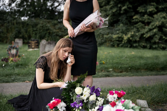 Woman crying at gravesite with flowers, another woman standing holding bouquet Woman crying at gravesite with flowers, another woman standing holding bouquet