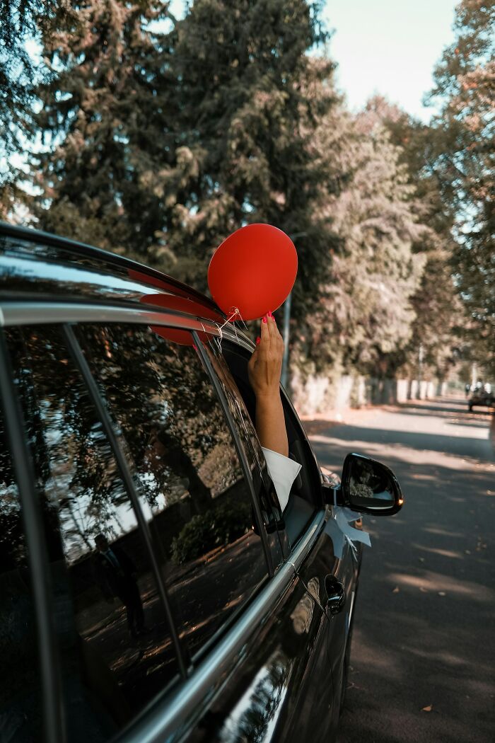 Person holding red balloon out of car window on tree-lined road