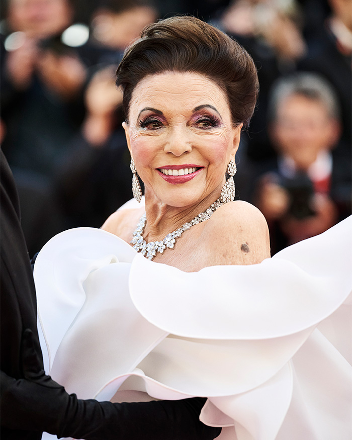 Close de Joan Collins sorrindo em um vestido branco com babados e colar de diamantes