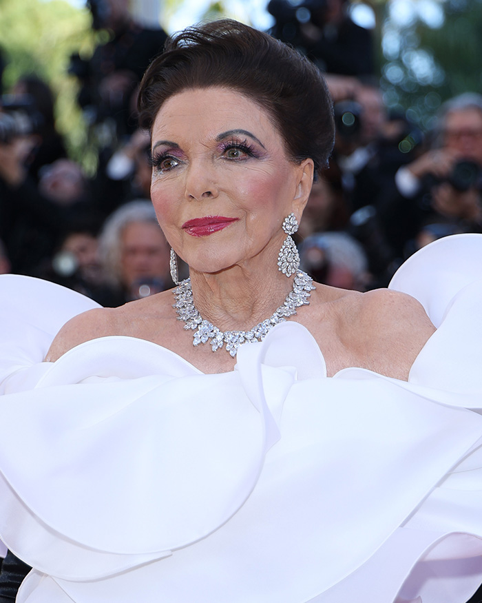 Joan Collins usando vestido branco com babados e joias de diamantes posando no tapete vermelho