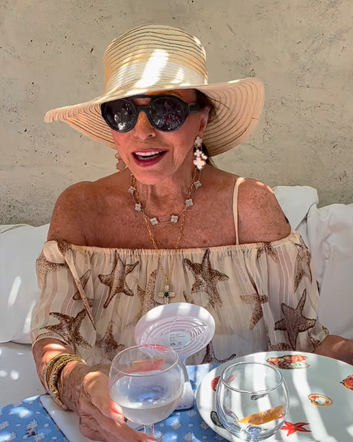 Joan Collins usando chapéu de sol e óculos escuros em mesa ao ar livre com bebidas
