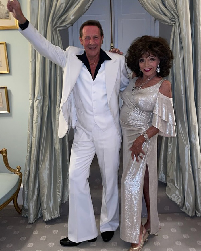 Joan Collins posando com um homem vestindo terno branco, ambos sorrindo dentro de casa