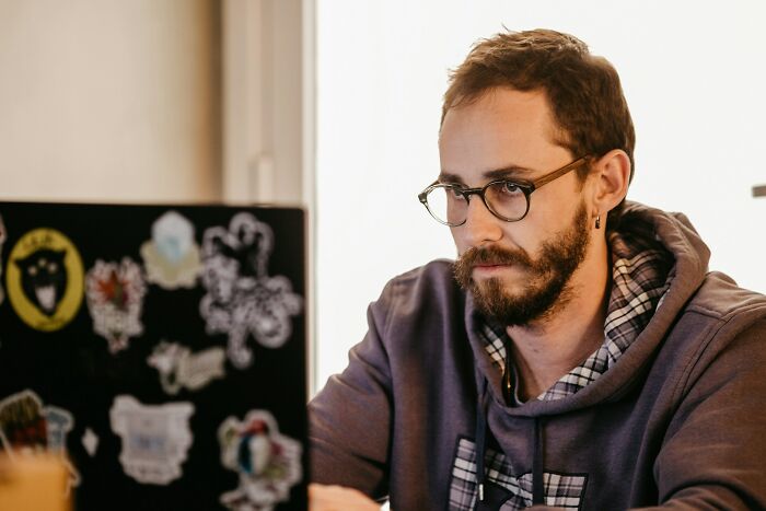A man with glasses and a beard, wearing a hoodie, intently looking at his laptop, exploring manosphere content.