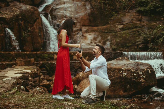 A man proposing to a woman in a red dress by a waterfall. Engagement stories of romance and chaos.