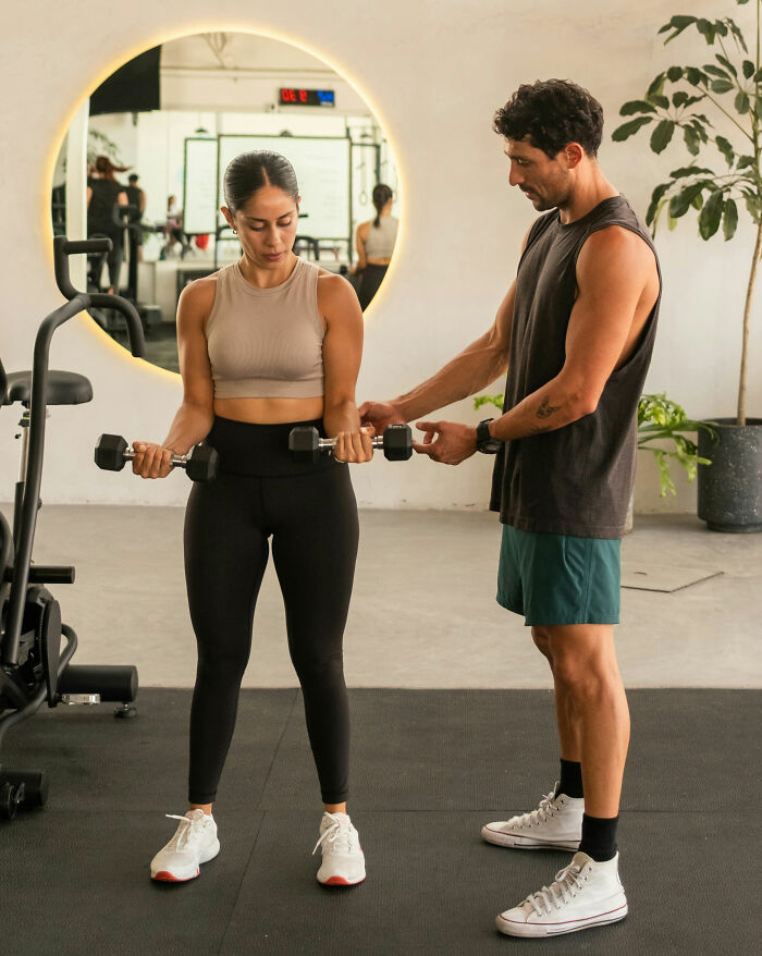 Woman lifting dumbbells with a trainer in a gym, demonstrating expensive habits that save you money.