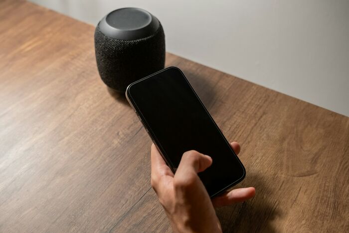 A person's hand using a smartphone next to a smart speaker on a wooden table, relating to smart scale cheating stories.