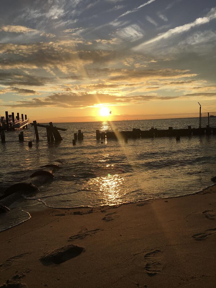 Sunset over water with footprints on sandy beach and old wooden pier remains