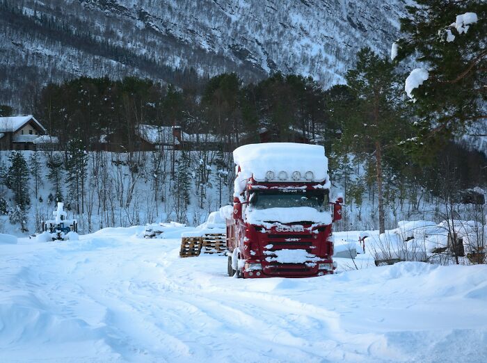 Snow-covered red truck parked in snowy mountainous forest area