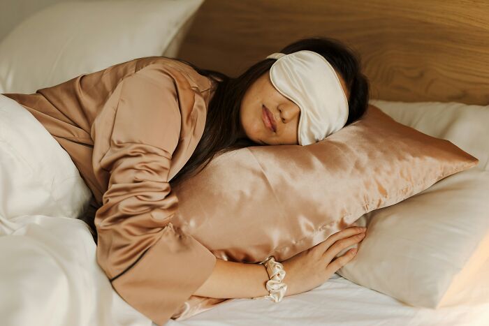 Woman sleeping soundly in bed with a silk eye mask and pillow, emphasizing habits that save money.