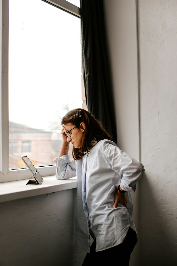 A woman looking out a window with a pained expression, hand on her back, suggesting the stress of wedding planning.