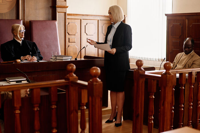 A lawyer addresses the court, holding papers, with a judge and defendant in Hard Cases, a courtroom scene.