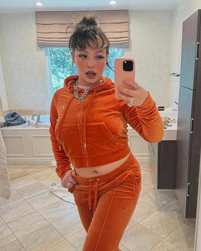 Lola Young poses in an orange velvet tracksuit, holding a phone for a mirror selfie, revealing how she caught cheating.