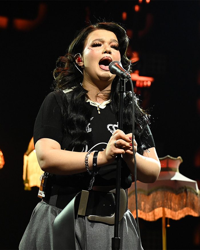 Lola Young singing into a microphone on stage, wearing a black top and gray skirt.
