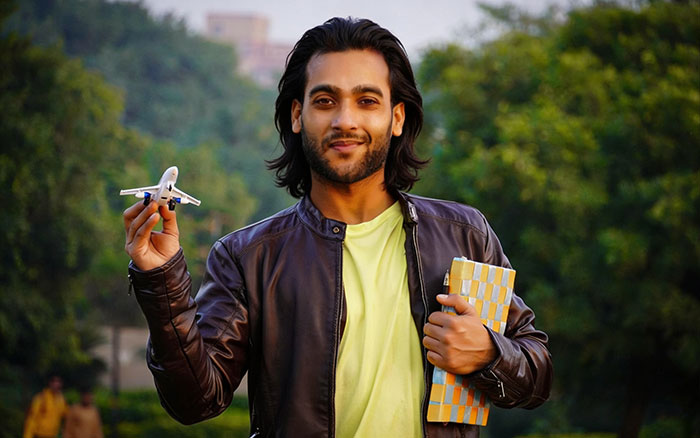 A smiling man in a leather jacket holds a model airplane and a book outdoors, perhaps a pilot.